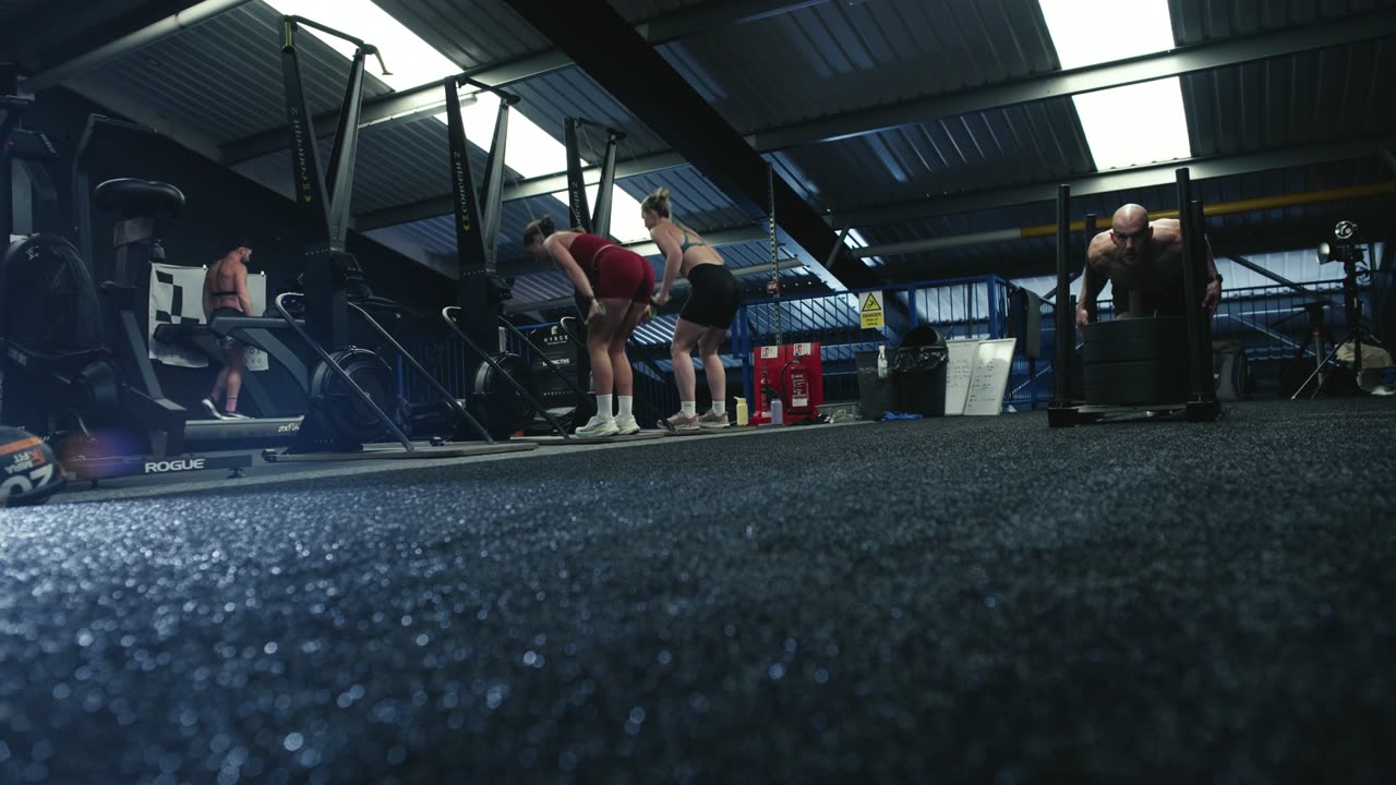 Gym floor during a HYROX sim: athletes at the SkiErgs while another pushes a sled