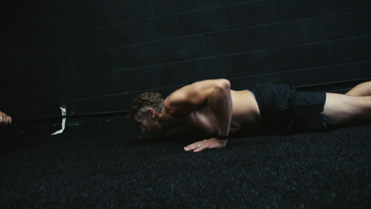Athlete mid-burpee on a dark gym floor during a competition